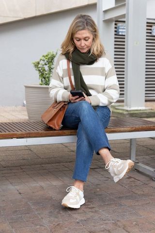 Middle-aged woman using smartphone on urban bench, casual outfit with leather bag