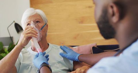 Caregiver Administering Oxygen Mask Comforting Elderly Patient in Home Care Living Room