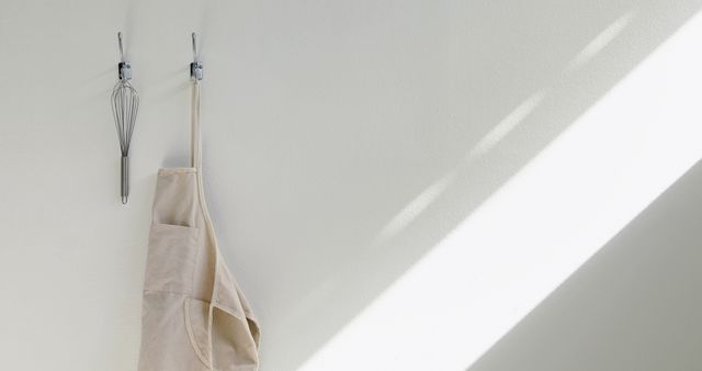 A whisk and an apron hang on a wall in a minimalist kitchen setting, with copy space. These items suggest a clean and organized cooking environment, ready for culinary activities.