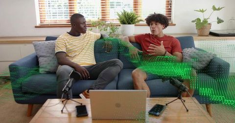 Two cohosts recording casual home podcast on couch with microphones and laptop