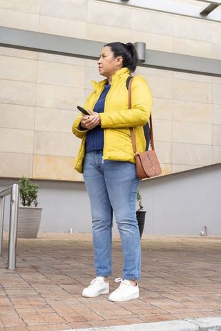 Mature woman standing with smartphone in yellow jacket on city forecourt, urban lifestyle