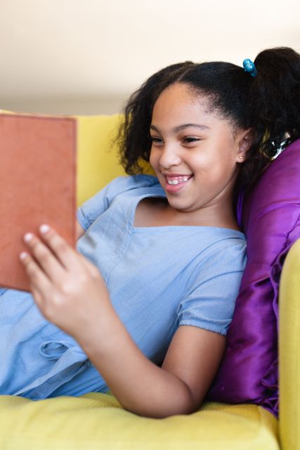 A cheerful biracial elementary schoolgirl is reading a book while sitting on a yellow couch with a purple pillow. She is smiling and appears to be enjoying her book. This image can be used for educational materials, children's books, school promotions, or articles about childhood learning and relaxation.