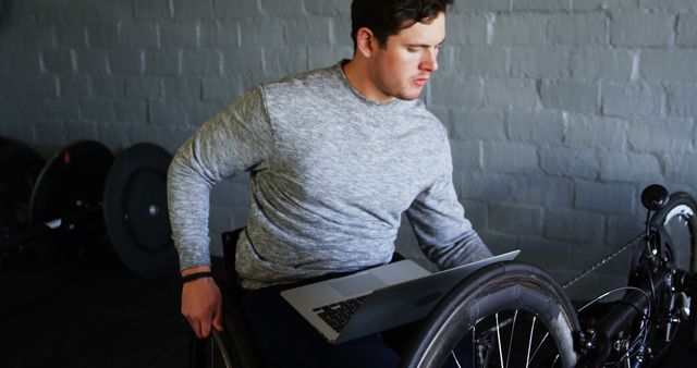 Person seated in a wheelchair using a laptop in a gym environment. Ideal for topics on disability inclusion, adaptive athletes, remote work, perseverance, and sports facilities.