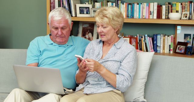 Elderly couple sitting on sofa, engaging with technology. The man uses a laptop while the woman holds a smartphone. This image can be used for topics related to seniors using technology, modern communication, aging, tech support, online activities for the elderly, and family bonding. The relaxed setting with bookshelves in the background adds a homey feel, making it suitable for lifestyle, retirement planning, and telecommunication content.