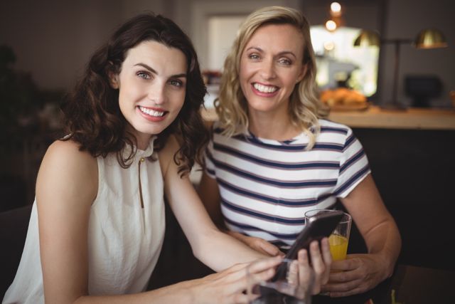 Two women are sitting in a restaurant, smiling and using a mobile phone. One woman is holding a drink. This image can be used for themes related to friendship, socializing, technology, and casual dining. It is suitable for advertisements, blog posts, and social media content promoting restaurants, mobile apps, or lifestyle topics.