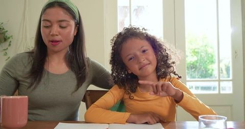Mother helping daughter with homework at sunlit table, child pointing to notebook