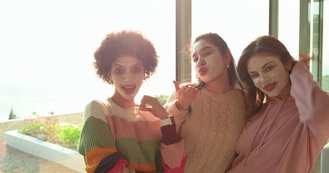 Multicultural friends enjoying sunlit pamper night on balcony with face masks and knitwear