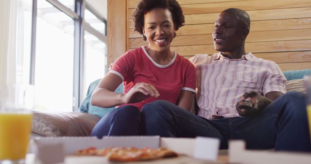 Image of happyafrican american couple sitting on sofa and eating pizza. Love, relationship and spending quality time together concept.