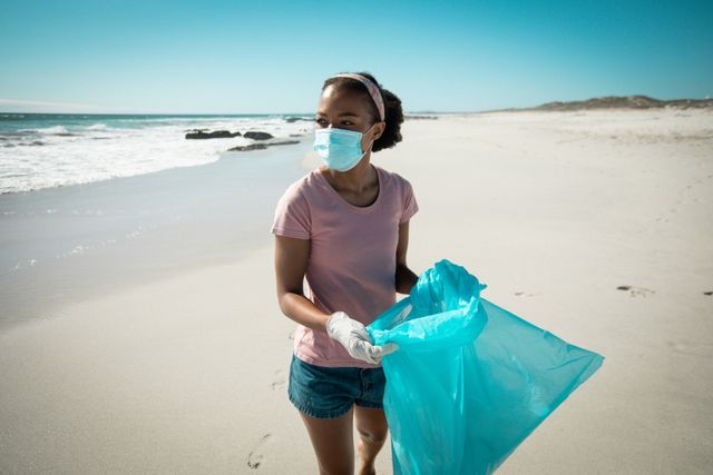 African american woman wearing face mask and gloves putting rubbish in refuse sack on sunny beach. eco conservation volunteer, beach clean-up.
