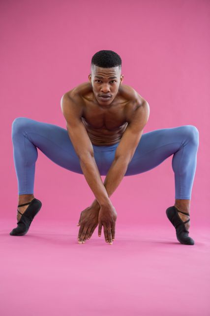 Male ballet dancer practising in studio against pink background. Wearing blue tights and black ballet shoes, demonstrating flexibility and strength. Ideal for use in articles about dance, fitness, performing arts, and professional training.