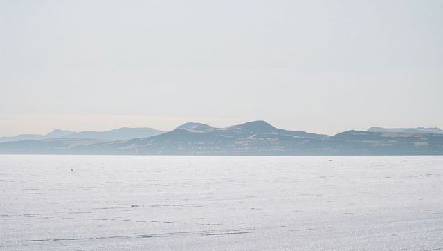 Minimalist monochrome frozen plain with distant mountain ridge and tracks under pale sky