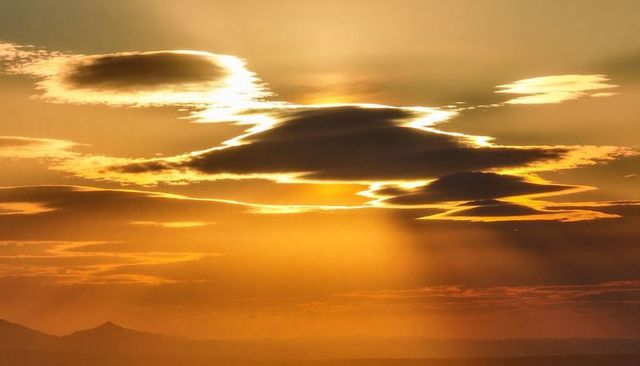 Golden lenticular clouds casting sunbeams over distant mountain ridge at sunset