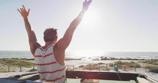 A man raises his arms in excitement while standing in a convertible near a beach, basking in sunlight with the ocean in the background. Suitable for themes of travel, adventure, freedom, and summer joy in promotional material, travel blogs, or ads.