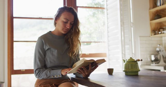 Happy caucasian woman sitting in cottage kitchen reading book, with teapot on counter - Download Free Stock Photos Pikwizard.com