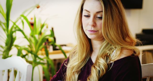 Young woman with long blond hair is deep in thought. Sitting indoors at home, she appears relaxed and casual. Plant life in background adds a peaceful atmosphere. Ideal for mental wellness, introspection, or home lifestyle themes.
