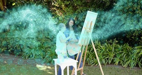 Senior chinese woman painting in garden with helper guiding, teal glow, outdoor art lesson