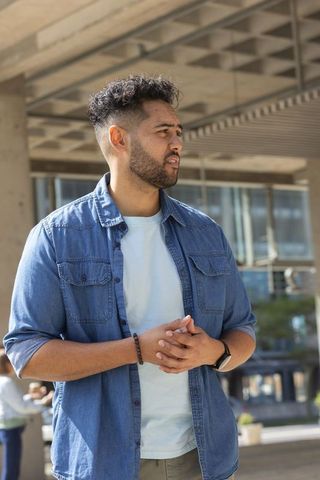 Standing man clasping hands beneath concrete canopy in urban plaza wearing denim shirt