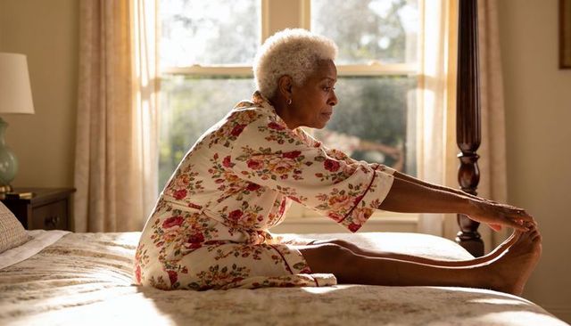 Senior woman stretching on bed in sunlit bedroom wearing floral robe for morning calm