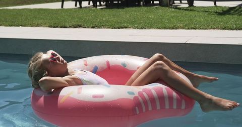 Young girl reclining on pink donut float in pool wearing red heart sunglasses, summer backyard