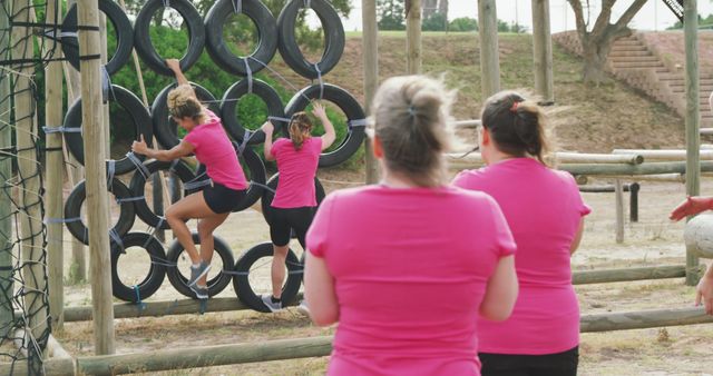 Caucasian female friends in pink t shirts watching and climbing tyres on climbing frame at bootcamp. Female fitness, challenge and healthy lifestyle.