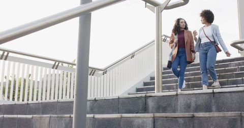 Two women walking down city stairs talking and carrying crossbody bags on overpass
