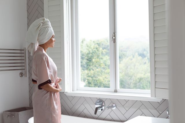 Woman standing in bathroom with towel on head, tying nightwear while looking out window. Ideal for themes of morning routine, self-care, relaxation, home life, and personal hygiene.