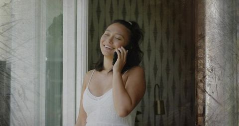 Smiling chinese woman talking on smartphone by sliding glass door in white dress, balcony