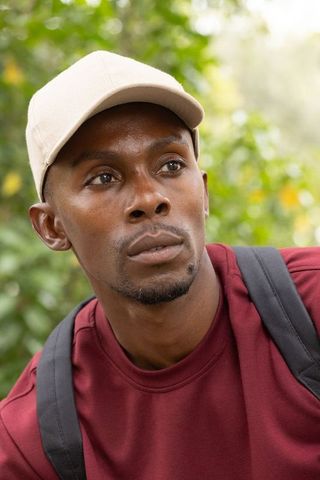 African american man wearing beige cap and maroon crewneck looking contemplative in leafy park