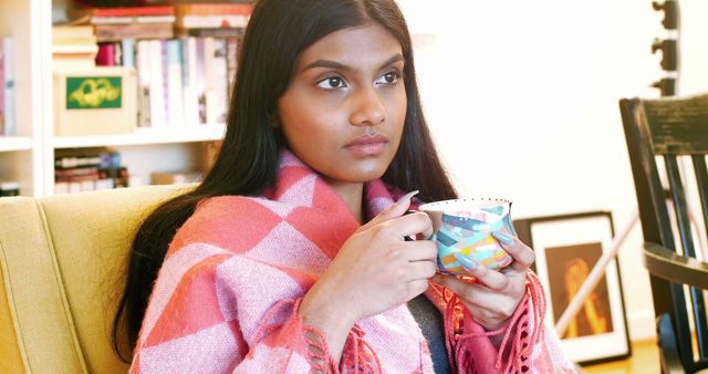 Young woman sitting on chair wrapped in blanket, holding colorful cup of tea. She appears deep in thought. Great for content about relaxation, self-care, lifestyle, and cozy home environments.