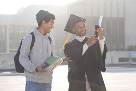 Sunlit diverse graduates celebrating on campus taking selfie with diploma and gown