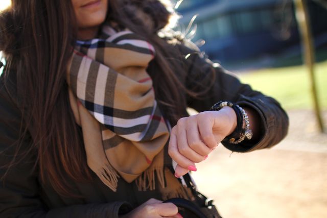 Woman wearing patterned scarf and jacket, looking at wristwatch in outdoor setting with sunlight. Suitable for themes related to lifestyle, fashion, time management, and outdoor activities.