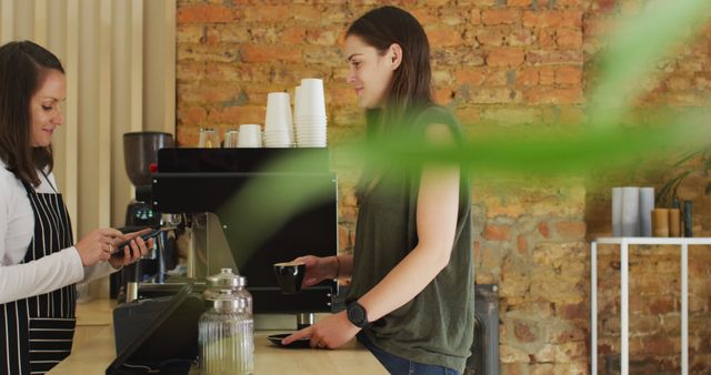 Scene depicting a friendly interaction between a barista and a customer in a rustic café. Customer holding a cup of coffee while the barista operates a handheld device, likely processing a payment. Ideal for use in contexts highlighting small businesses, cozy coffee shop atmospheres, or casual urban dining experiences.