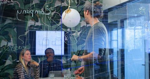 Presenter writing on glass partition during collaborative team meeting with floorplans