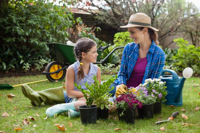 A mother and daughter are sitting on the grass in their backyard, surrounded by colorful flowering potted plants. They are smiling and enjoying time together while gardening. This scene can be used for themes related to family bonding, outdoor activities, gardening, and nature. Ideal for promotional materials, social media content, blogs, and articles about family life, nature conservation, and hobbies.