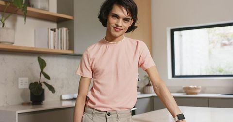 Young man wearing pale-pink shirt and pearl necklace leaning on kitchen countertop with smartwatch
