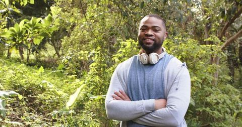 African american man smiling in sunlit forest wearing headphones and hoodie, arms crossed