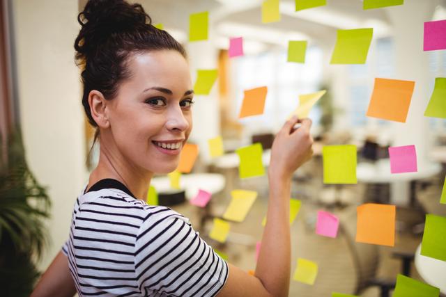 Businesswoman smiling while organizing colorful sticky notes on glass in a modern office. Ideal for use in articles or advertisements about office productivity, teamwork, project management, and creative brainstorming sessions.