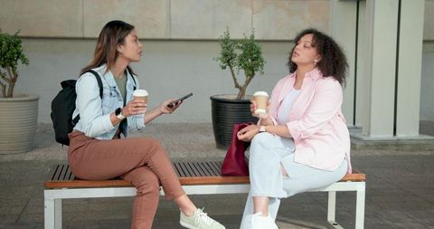 Diverse young women chatting over coffee on urban bench holding smartphone and cups