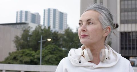 Mature woman wearing headphones and hoodie standing on city rooftop, contemplating skyline
