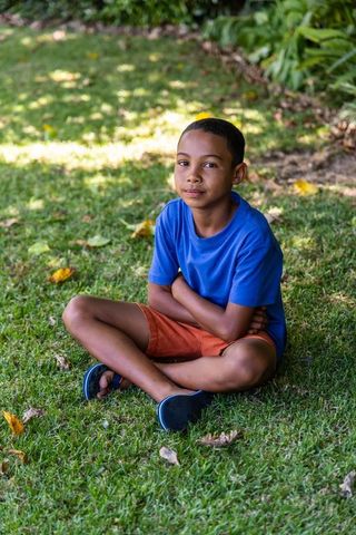 African preteen boy sitting on grass with arms folded wearing blue tee and orange shorts