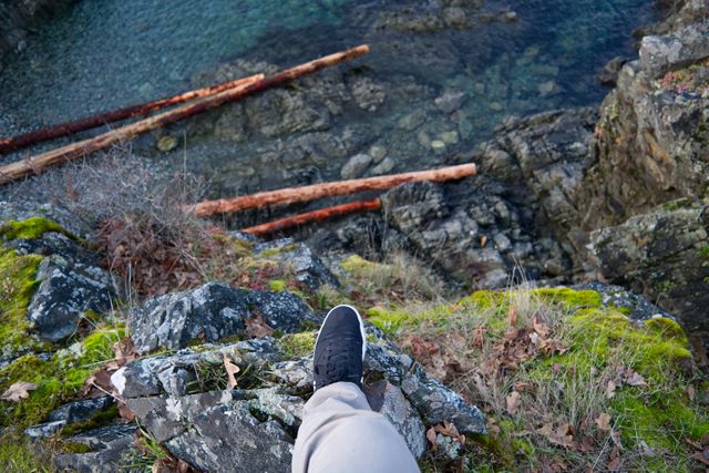 Person is standing on a rocky cliff edge covered with moss and fallen leaves, looking down at crystal clear water and rocks below. Perfect for themes of outdoor adventure, nature exploration, and travel. Can be used in websites, blogs, or advertisements focusing on hiking, travel destinations, and thrilling experiences.