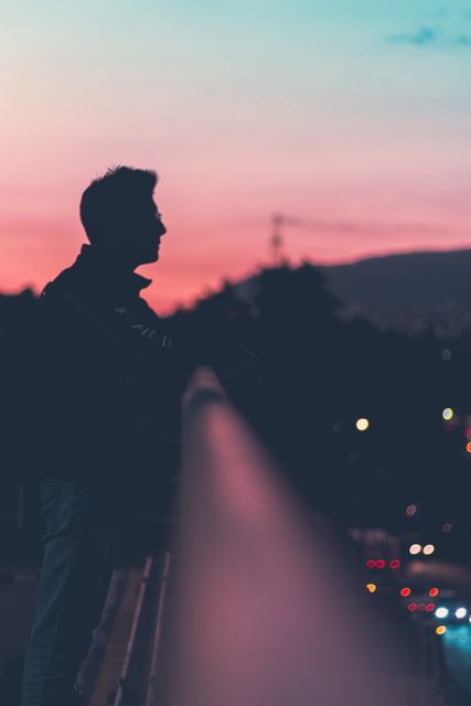 This image portrays a silhouetted man standing on a bridge against a beautiful sunset sky. Ideal for themes of contemplation, relaxation, and urban tranquility. Suitable for use in blogs, articles, or social media posts about finding peace in the city, mindfulness, or evening reflections.