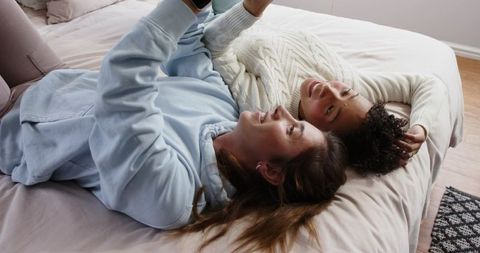 Diverse mid-adult women lying on bed sharing smartphone, cozy casual bedroom moment