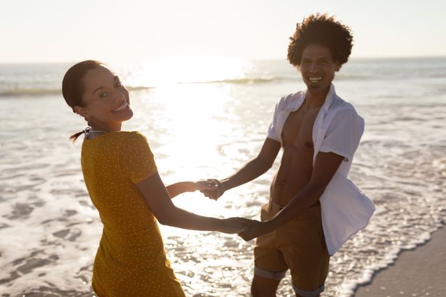 Young couple holding hands and smiling while enjoying a beautiful sunset at the beach. Perfect for travel brochures, romantic getaway promotions, summer vacation ads, and lifestyle blogs focusing on relationships and leisure activities.
