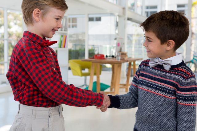 Two boys dressed in formal attire shaking hands in a modern office environment. This image can be used to illustrate concepts of teamwork, cooperation, young entrepreneurship, and childhood friendships in a professional setting.