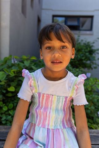 Young girl sitting on bench in courtyard wearing pastel striped dress and white top