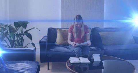 Young woman reading papers on dark blue sofa with tablet and notebook in modern living room