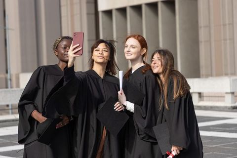 Multicultural women graduates taking selfie on campus wearing black gowns holding diplomas