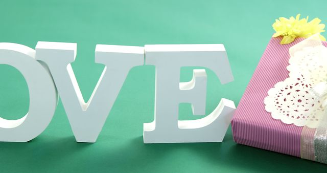 Large white letters spelling LOVE are set against a green background, with a pink gift box adorned with a yellow flower and white lace to the side, with copy space. It conveys a theme of affection and celebration, for occasions like Valentine's Day or anniversaries.