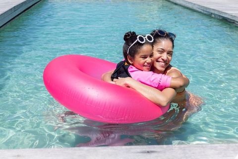 African american mother hugging daughter on pink inflatable ring in sunny outdoor pool